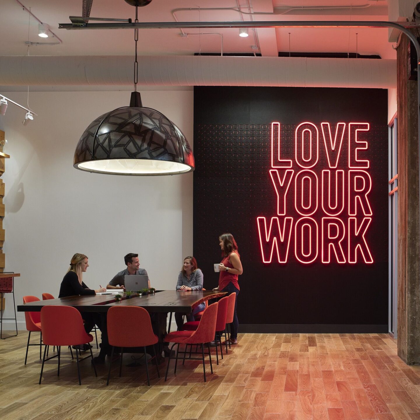 Four people work together at a table infront of a neon sign that reads "love your work"